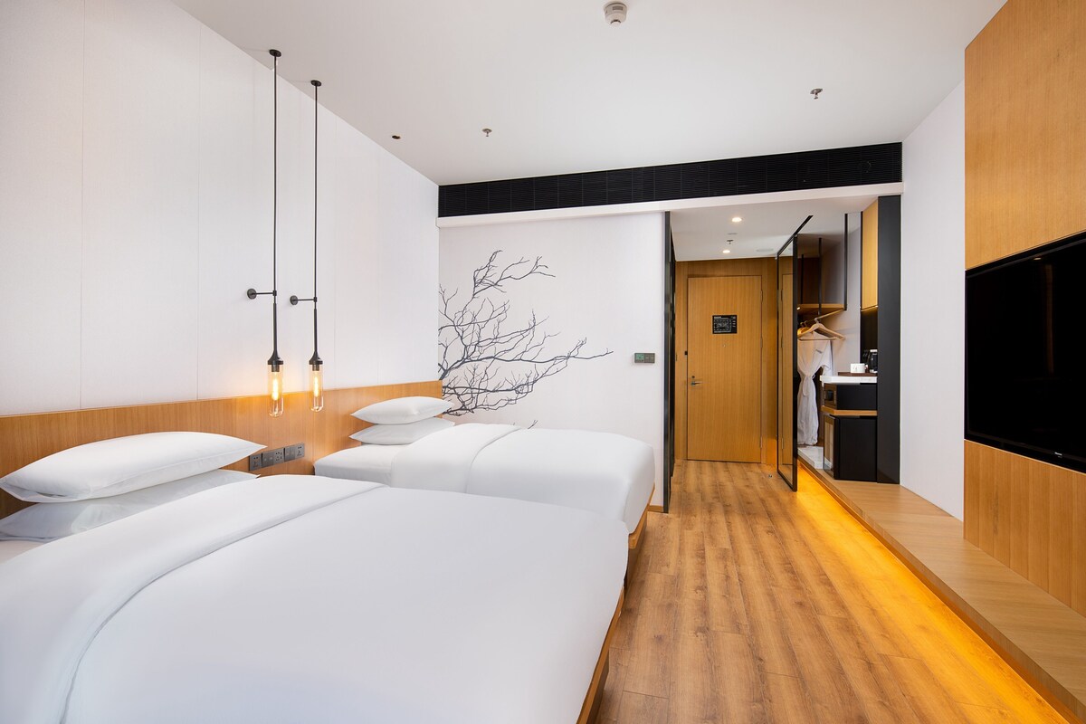 A modern hotel room with two beds, a wooden floor, a wall-mounted TV, and a hallway leading to a bathroom.