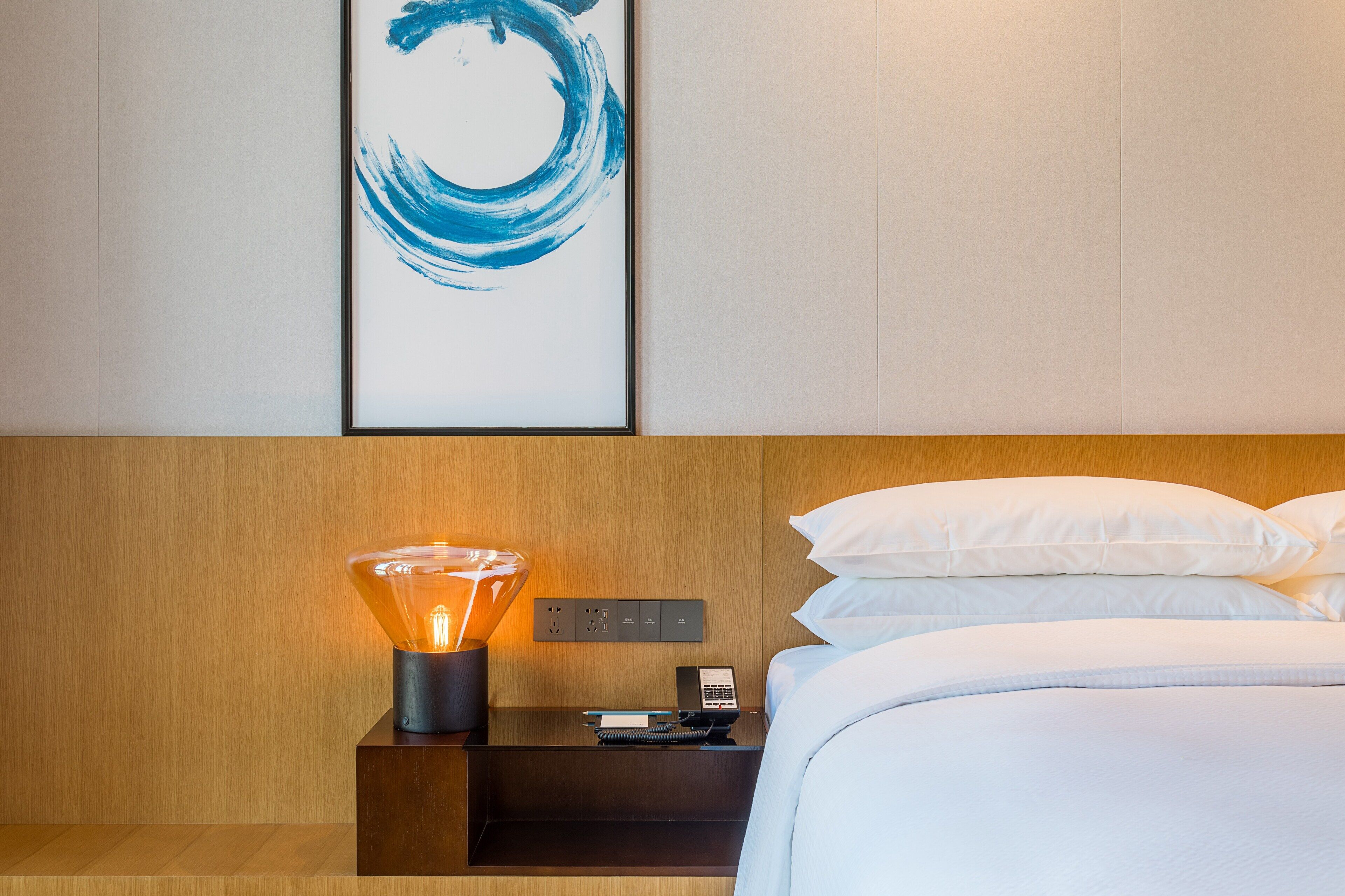A neatly made bed with white linens, a bedside table with a lamp, a phone, and a power outlet.