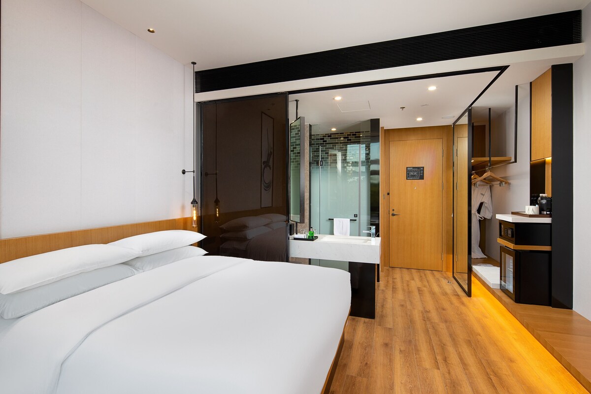 A modern hotel room with a large bed, a wooden floor, a bathroom with a glass shower, and a small kitchenette.
