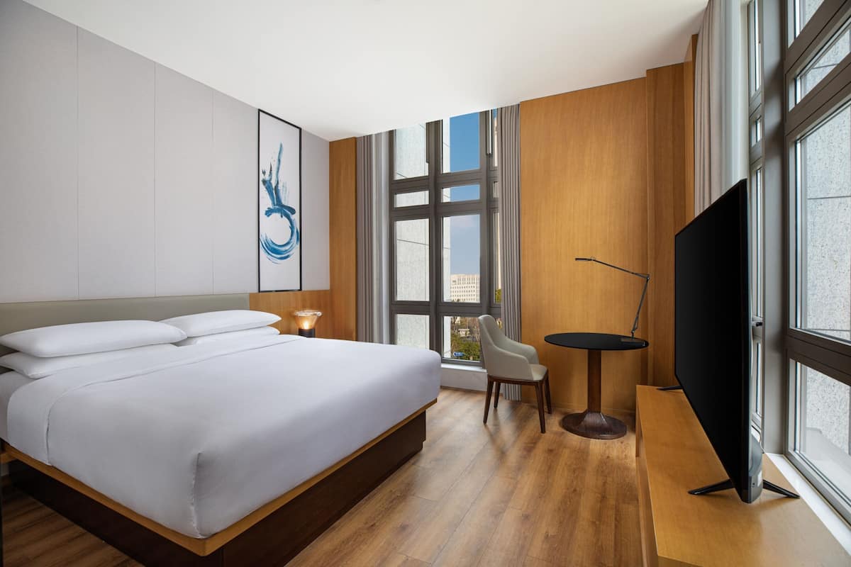 A modern hotel room with a large bed, a desk with a chair, a television, and a large window offering a city view.
