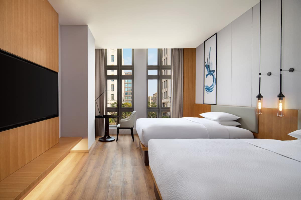 A modern hotel room with two beds, a desk, and a large window offering a city view.