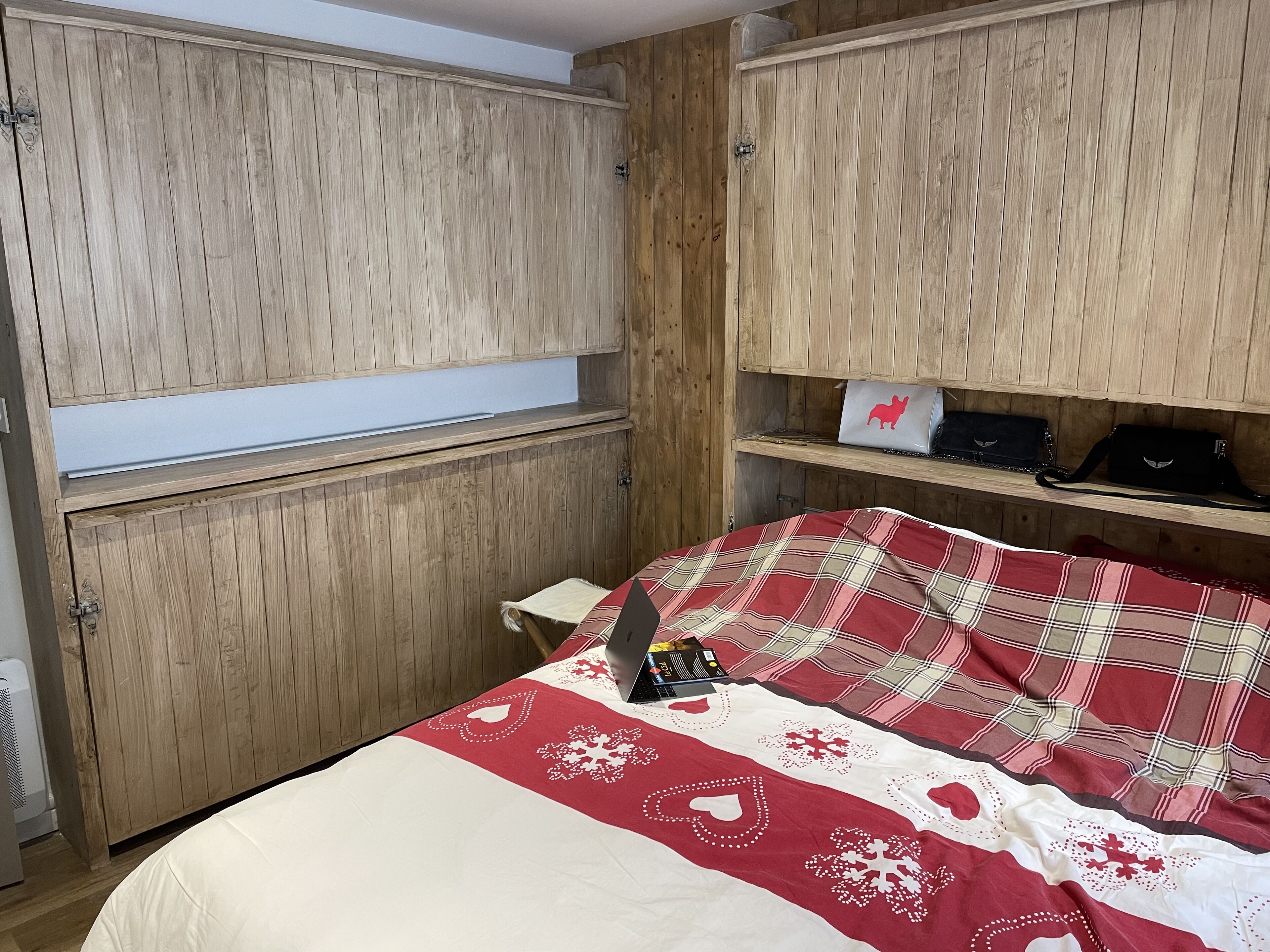 Une chambre avec des murs recouverts de panneaux de bois, un lit avec une couverture à carreaux rouges et blancs, et une étagère avec des sacs et un ordinateur portable.