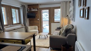 Ein gemütliches Wohnzimmer mit einer Couch, einem Couchtisch und einem Essbereich mit Tisch und Stühlen. Ein Kamin, ein Fernseher und ein Blick auf eine verschneite Landschaft sind durch die Fenster zu sehen.