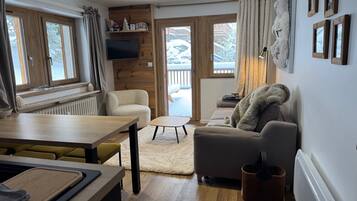 A cozy living room with a sofa, a coffee table, and a dining area with a table and chairs. There is a fireplace, a television, and a view of a snowy landscape through the windows.