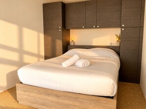 A neatly made bed with a rolled white towel, dark wooden headboard, and bedside lamps.