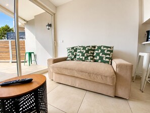A beige sofa with a patterned pillow, a remote control on a wooden table, and a black perforated stool. - Terrasse ensoleillée : appartement 1 chambre 1 cabine vue mer terrasse (Le Grau-du-Roi)