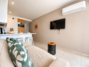 A modern living room with a sofa, a TV mounted on the wall, and a kitchen area with a microwave and oven. - Terrasse ensoleillée : appartement 1 chambre 1 cabine vue mer terrasse (Le Grau-du-Roi)
