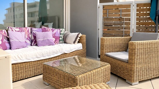 Un balcon aménagé avec des meubles en osier, notamment un canapé et des fauteuils, ainsi qu’une table basse en verre.