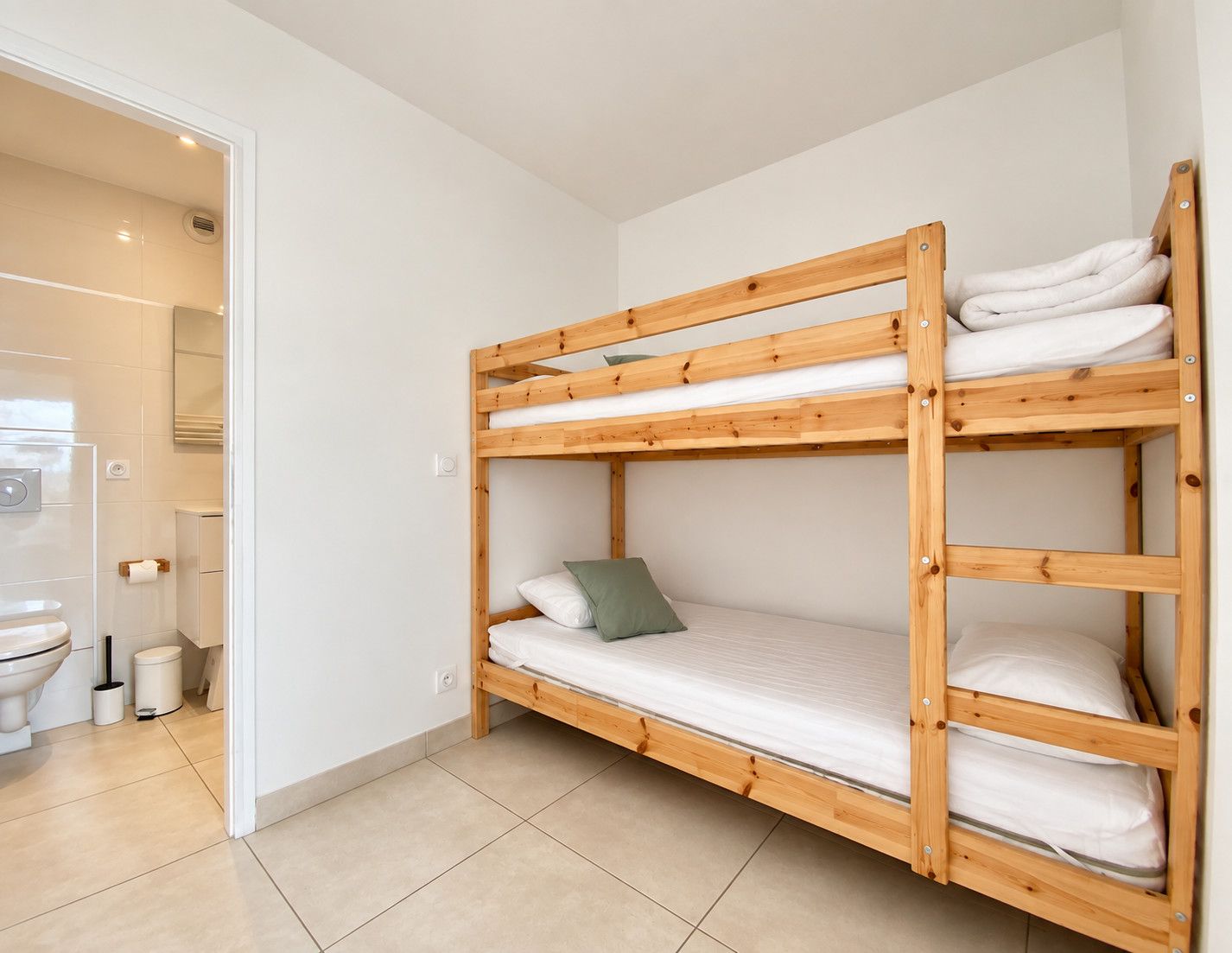 Una cama litera con estructura de madera, ropa de cama blanca y un cojín verde en una habitación con paredes blancas y suelo de baldosas.