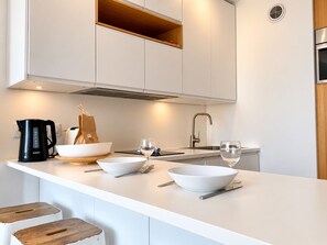 A modern kitchen with white cabinetry, a stainless steel range hood, a built-in oven, and a white countertop with bowls, a coffee maker, and a wine glass.