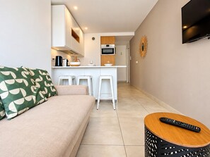 A modern living room with a sofa, a coffee table, and a kitchen area with a TV, bar stools, and a microwave.
