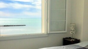 A bedroom with a large window offering a view of the sea. There is a bed with a grey and white quilt, a bedside table with a lamp, and a wall-mounted shelf.