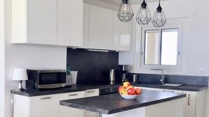 A modern kitchen with white cabinets, a black countertop, a central island with a stool, a microwave, a coffee maker, and a fruit bowl.