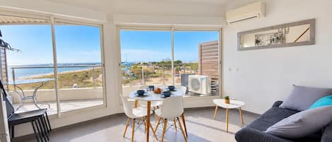 A modern living room with a dining area, large windows with a view of the sea, and a grey sofa.