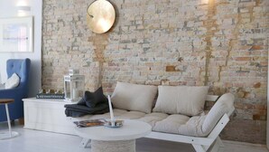 Una sala de estar moderna con pared de ladrillo, un sofá blanco, un sillón azul, una mesa de centro redonda y un aparador blanco con una lámpara y libros.