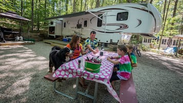 Una familia comiendo en una mesa de picnic con mantel de lunares, una parrilla y una caravana al fondo.