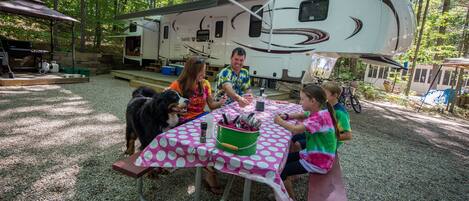 A family dining at a picnic table with a polka dot tablecloth, a grill, and a RV in the background.