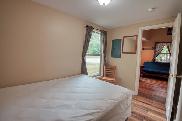 A bedroom with a large bed, a chair, a window with curtains, and a door leading to another room. - Danforth Bay Camping and RV Resort (Freedom)
