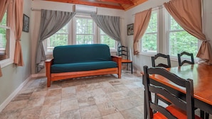 A cozy room with a wooden ceiling, a green sofa, a wooden dining table with chairs, and large windows with curtains.