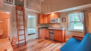 A compact kitchen with wooden cabinets, a microwave, and a dishwasher. There is a wooden dining table and chairs, a blue sofa, and a window with curtains. A wooden ladder is visible in the corner.