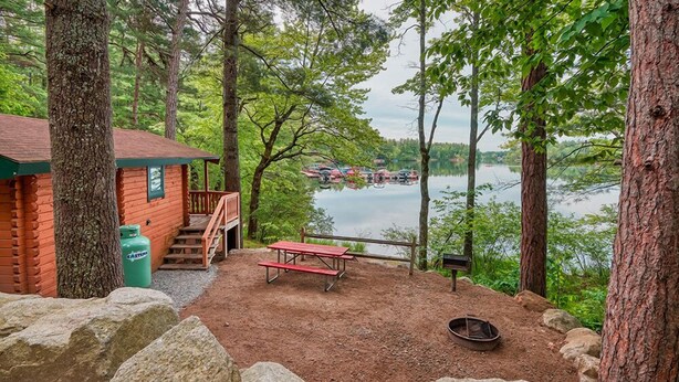 A cabin with a picnic table and a fire pit, surrounded by large rocks and trees, overlooking a lake with boats.