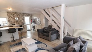 En moderne stue med en grå sofa, et hvidt sofabord og et gulvtæppe med et mønster. Et køkken med en vaskemaskine og en trappe, der fører op til en etage, er synligt.