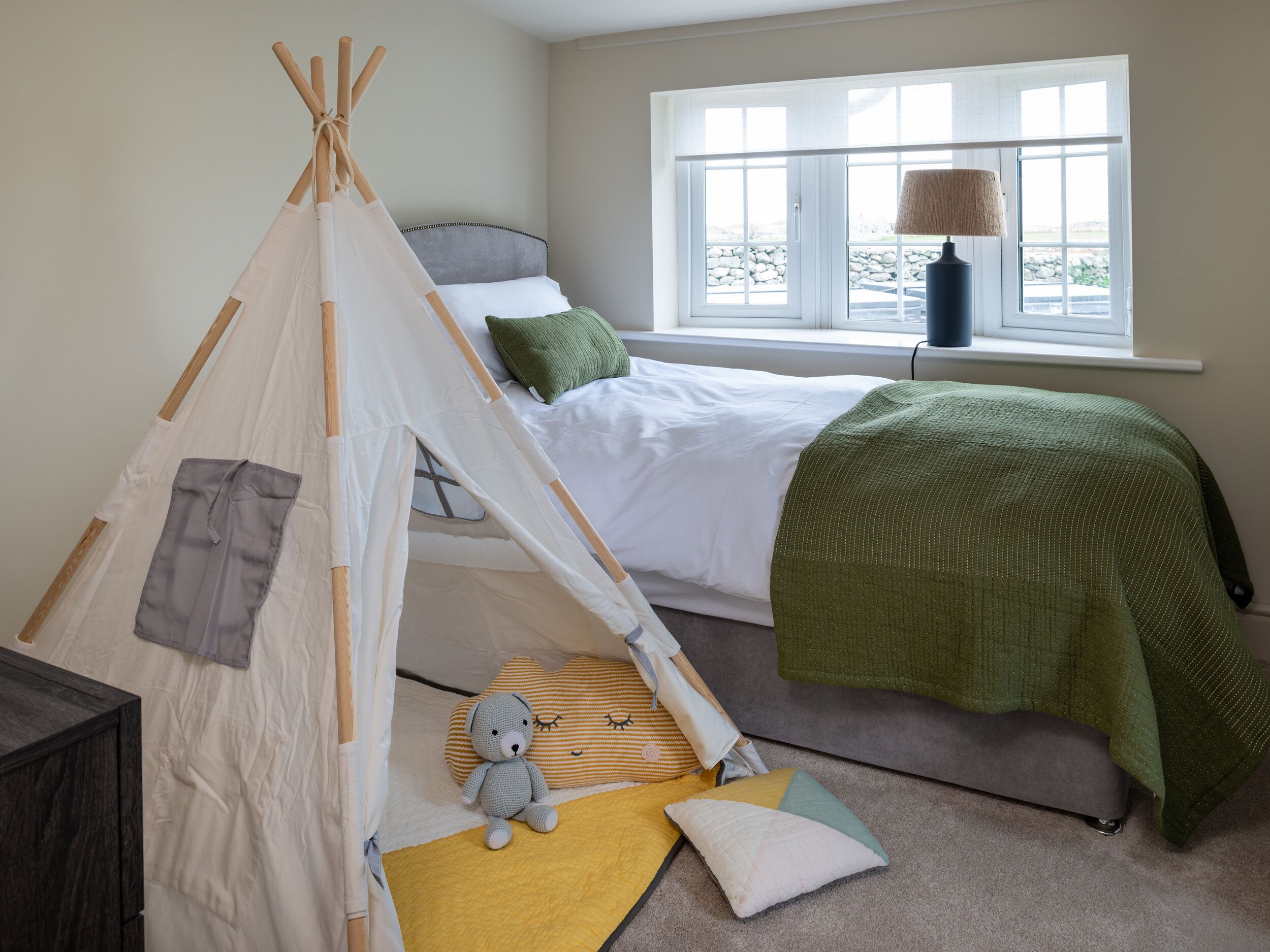 A bedroom with a bed, a green blanket, a small tent, a lamp, and a teddy bear.