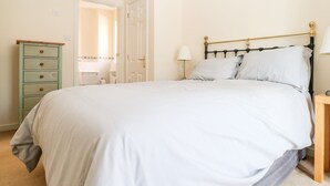 Een net opgemaakt bed met witte lakens, een groene dressoir met lades, een bedlampje en een deur die naar een andere kamer leidt.