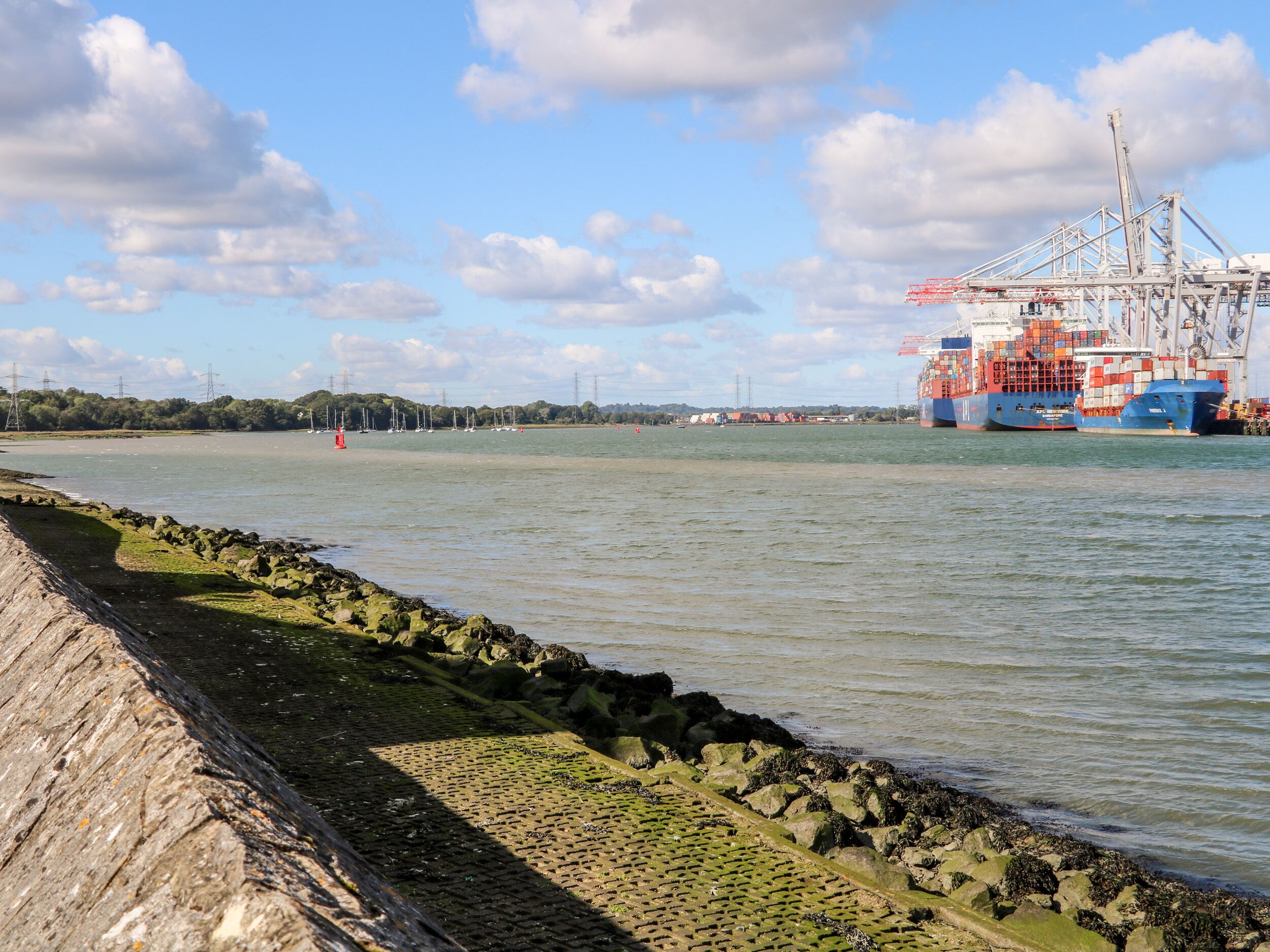 A harbor with a large cargo ship docked, a rocky breakwater, and a clear sky.