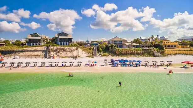 Un resort en la playa con aguas turquesas cristalinas, sombrillas y varios edificios al fondo.