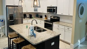 Una cocina con armarios blancos, un refrigerador de acero inoxidable, un microondas, una estufa y un lavamanos con encimera de granito.