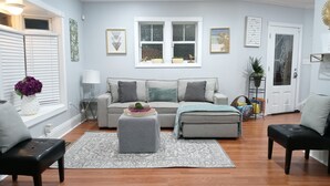 Una sala de estar con sofá, reposapiés y mesa de centro, una ventana con persianas y una puerta con un espejo decorativo.