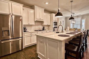 A modern kitchen with white cabinetry, stainless steel appliances, a central island with stools, pendant lighting, and a view of the outdoors.