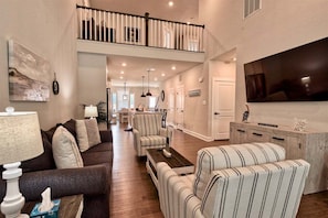 A spacious living room with a high ceiling, a staircase, a dining area, and a flat-screen TV mounted on the wall.