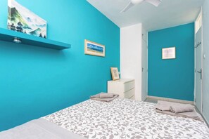 Une chambre avec des murs de couleur turquoise, un lit avec une literie à motifs, une commode blanche et un tableau encadré accroché au mur.