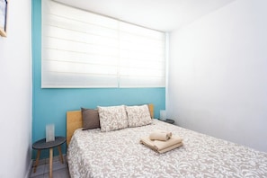 A bedroom with a bed, a nightstand, a window with blinds, and a wall with a blue accent.