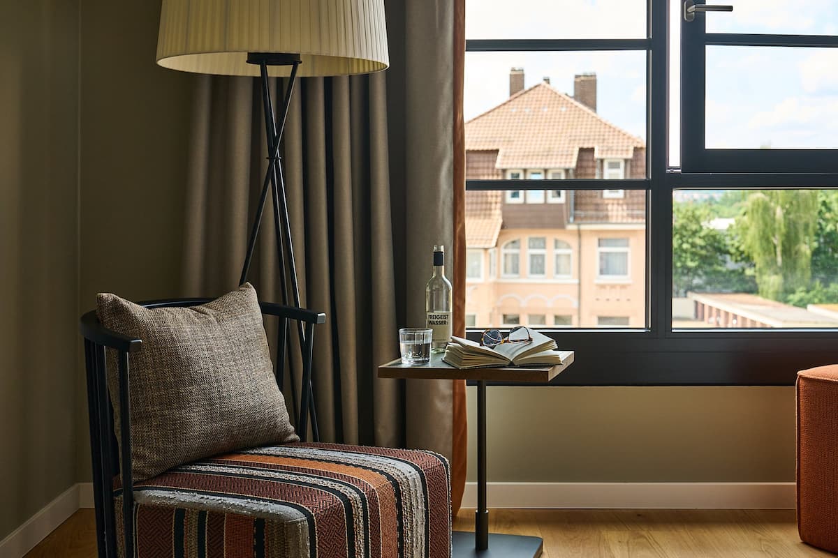 A cozy room with a chair, a table with a bottle, glasses, and an open book, a lamp, and a window with a view of buildings.