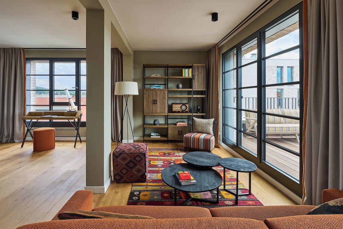 A modern living room with a large window, a bookshelf, a round coffee table, and a patterned rug.