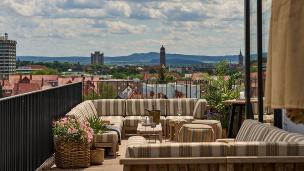 A rooftop terrace with a sofa set, a table with a vase, and a view of the cityscape including a tower. - FREIgeist Göttingen Nordstadt - A Member of Design Hotels™ (Goettingen)