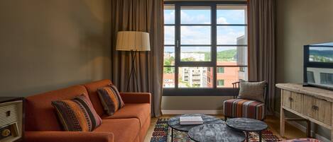 A modern hotel room with a sofa, armchair, and a small table. A large window offers a view of buildings and greenery.