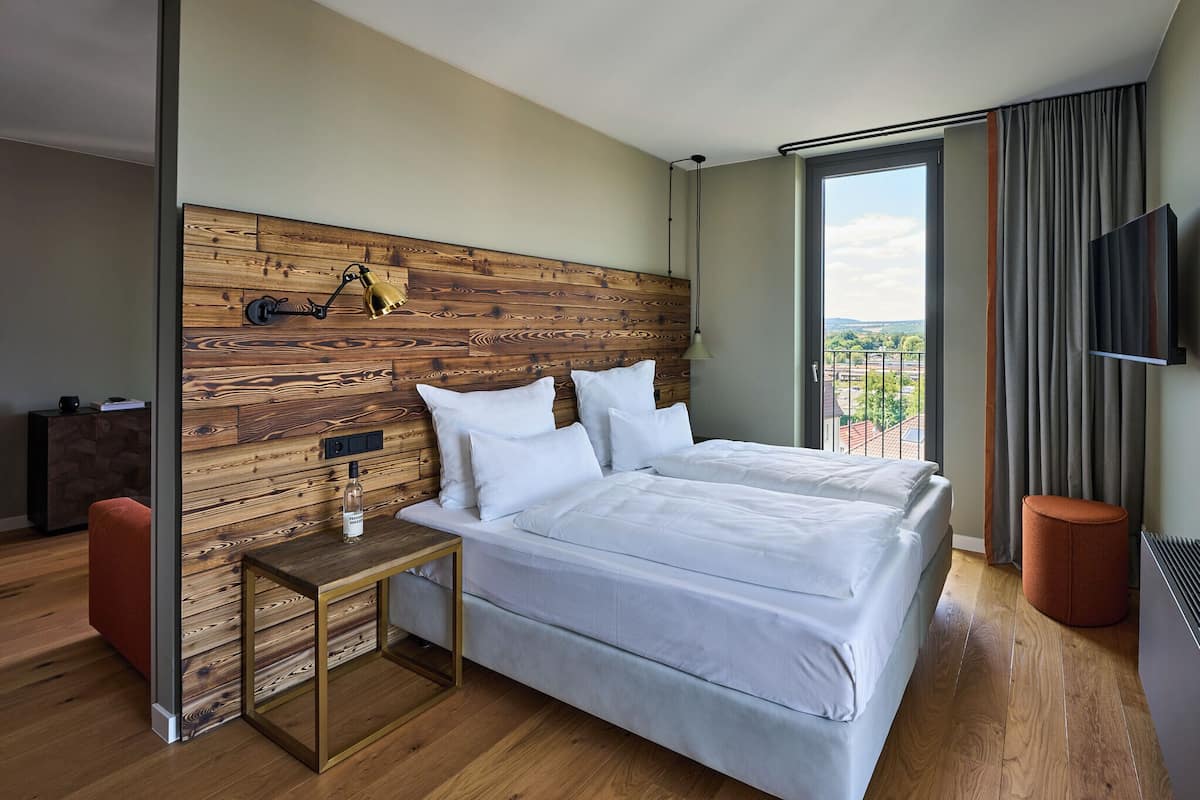 A hotel room with a wooden headboard, a bed with white bedding, a small table with a bottle, and a view of the outdoors.