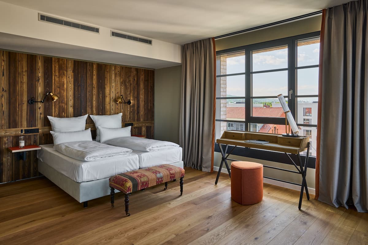 A bedroom with a wooden wall panel, a bed with two pillows, a bench, a desk with a chair, and a window with curtains.