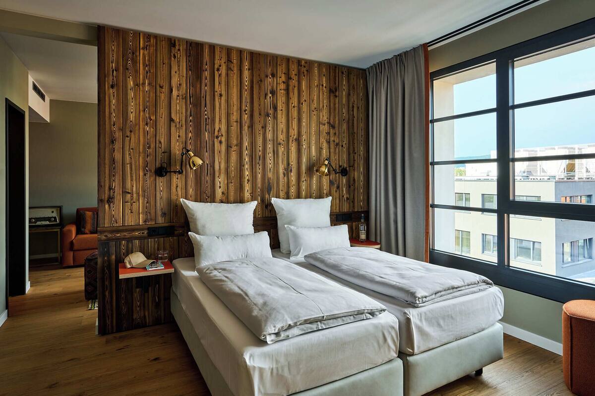 A double bed with a wooden headboard, a nightstand with a lamp, a window with a view of buildings, and a small orange chair.