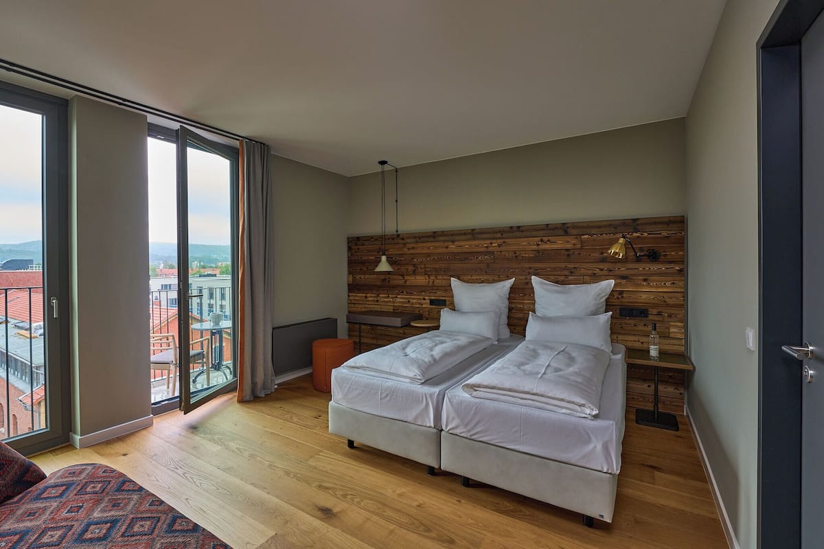 A modern bedroom with a large bed, a wooden headboard, a small table, and a view of the city through large windows.