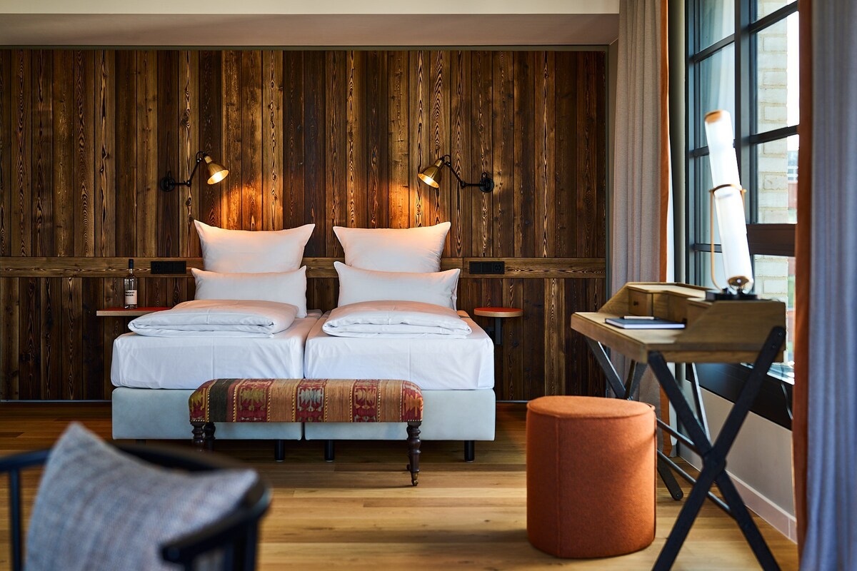 A double bed with white linens, two bedside tables with lamps, a wooden desk, and a large window with curtains.