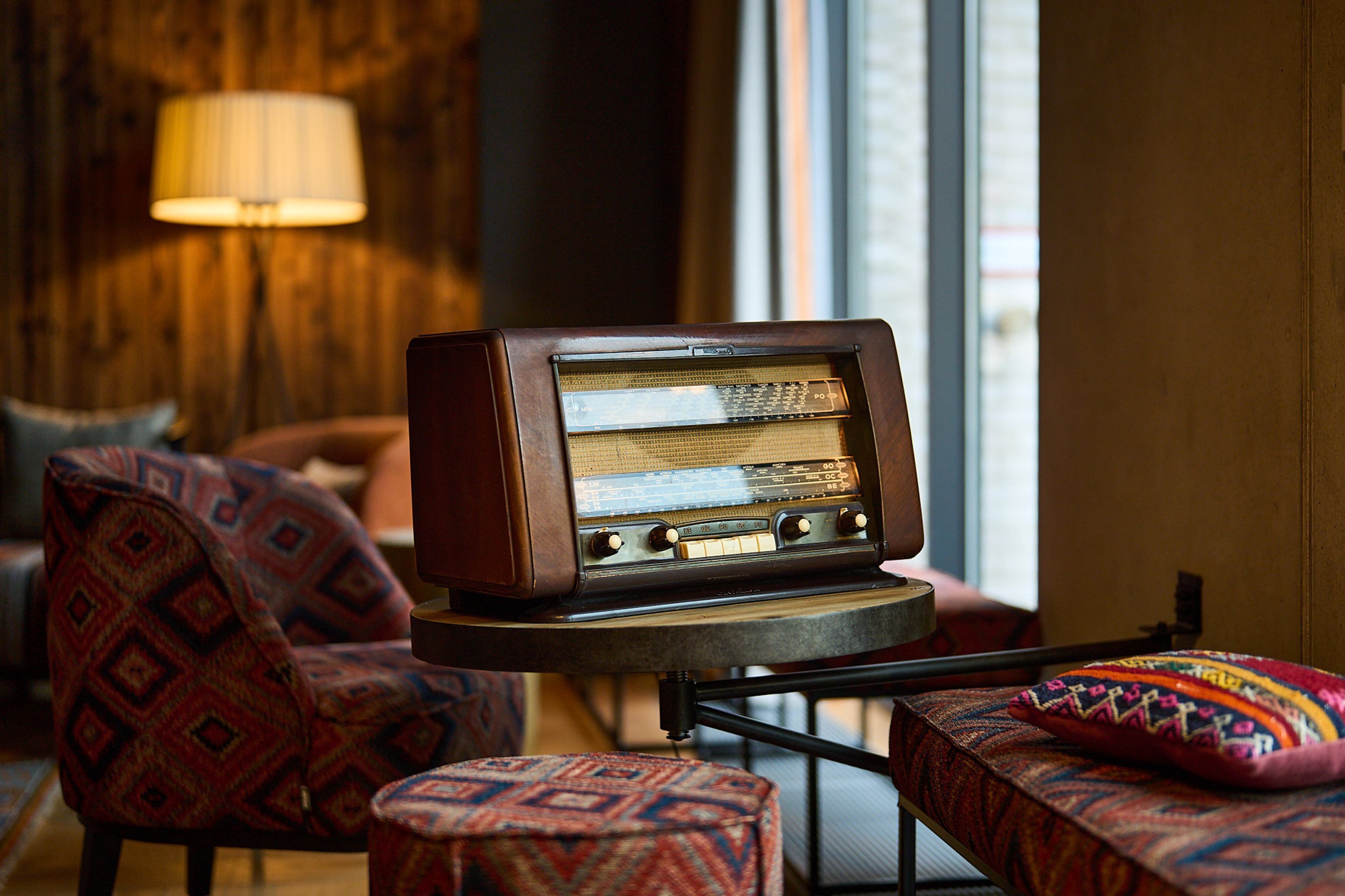 A vintage radio on a small table in a cozy room with patterned furniture and a lamp in the background.