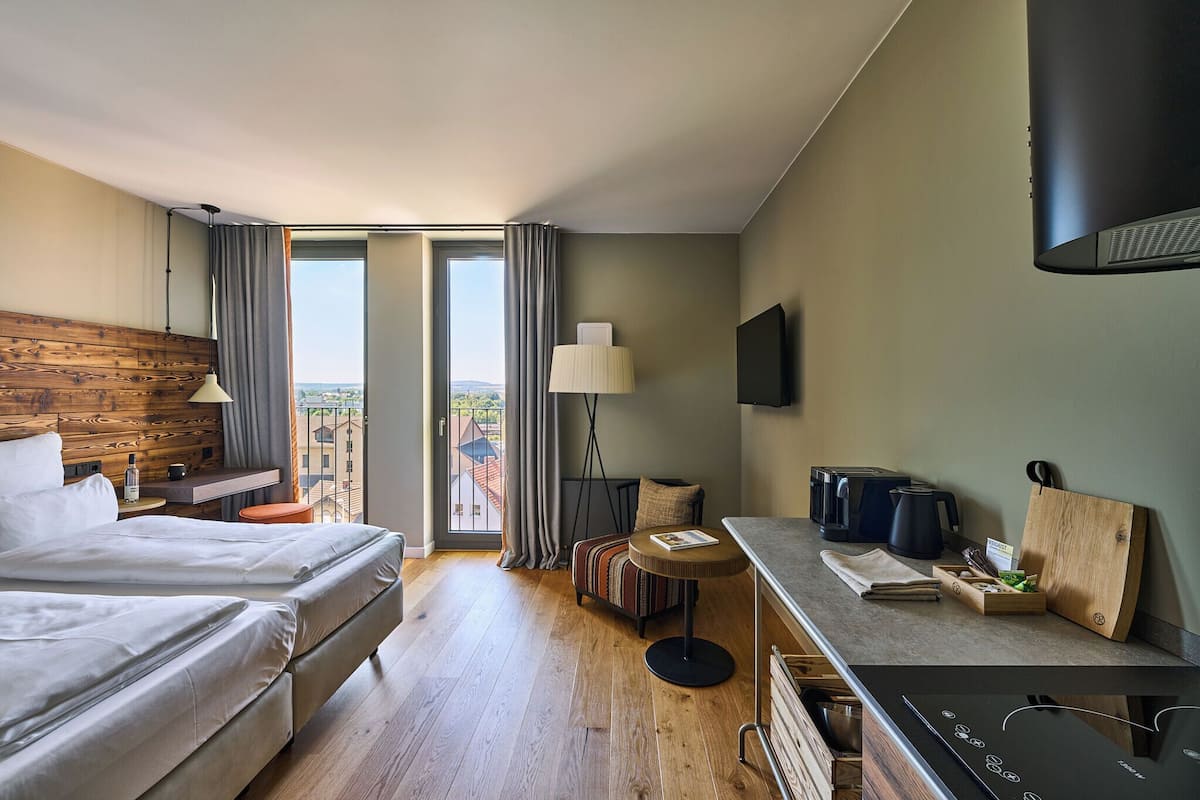 A hotel room with two beds, a dining area, a view through the window, and a flat-screen TV mounted on the wall.