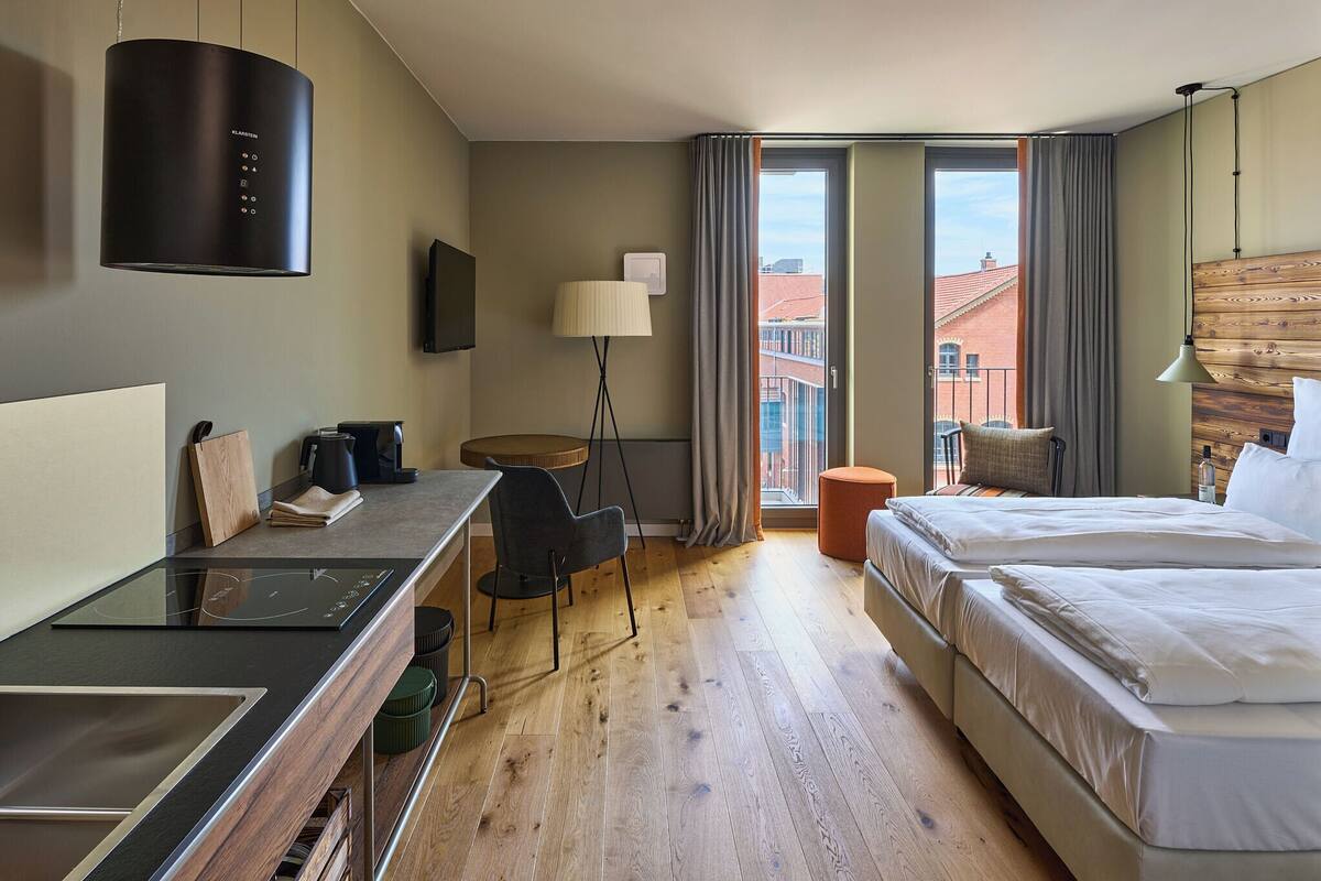 A hotel room with a bed, a desk with a chair, a standing lamp, a TV, and a view of buildings outside.