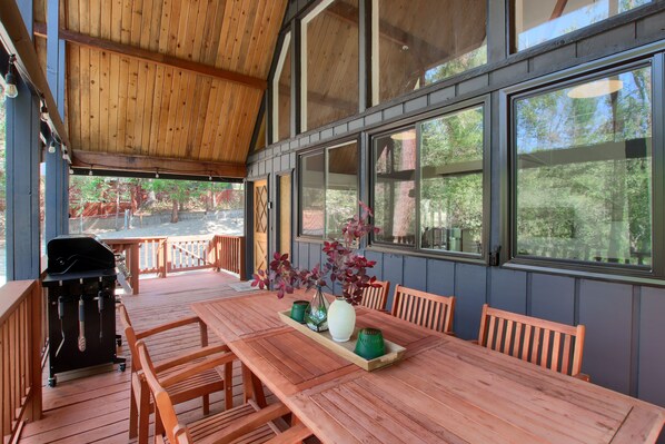A covered outdoor dining area with a wooden table and chairs, a grill, and large windows offering views of the surrounding trees. - Modern A-Frame next to a creek, minutes to Bass Lake and close to Yosemite (Oakhurst)