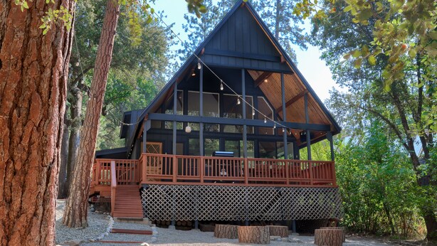 A cabin with a wooden deck, large windows, and a sloped roof, surrounded by trees. - Modern A-Frame next to a creek, minutes to Bass Lake and close to Yosemite  (Oakhurst)